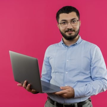 young bearded man in glasses and blue shirt holding laptop looking at camera smiling confident standing over pink background